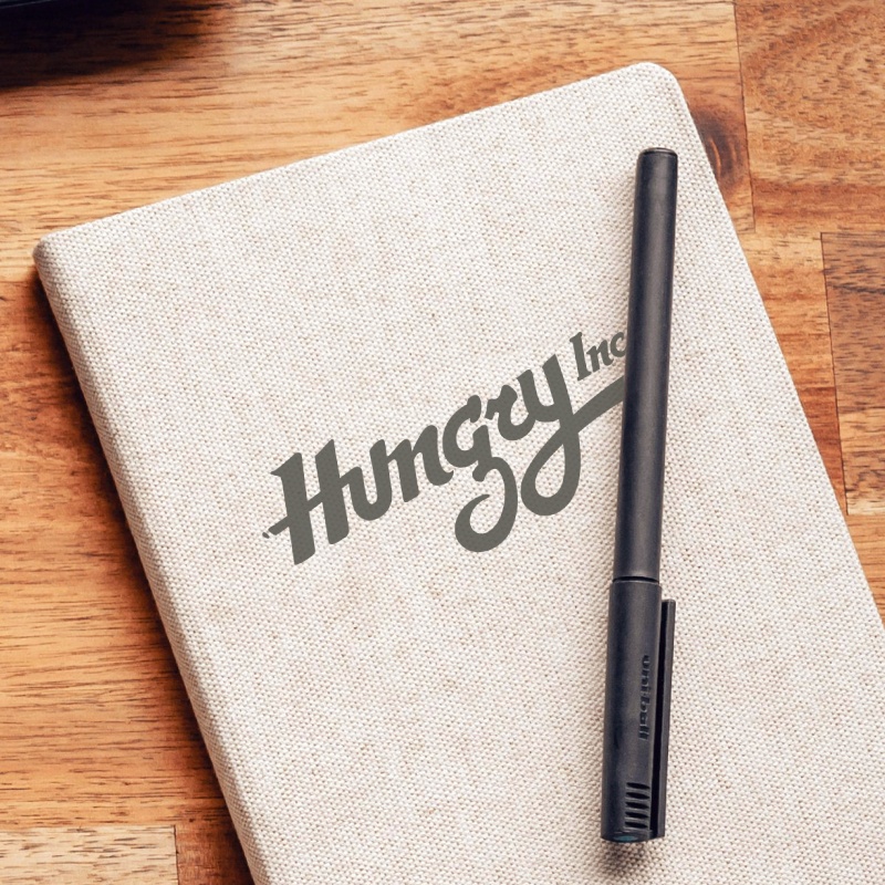 A paper notebook with a stamped lettering logo that says Hungry Inc.